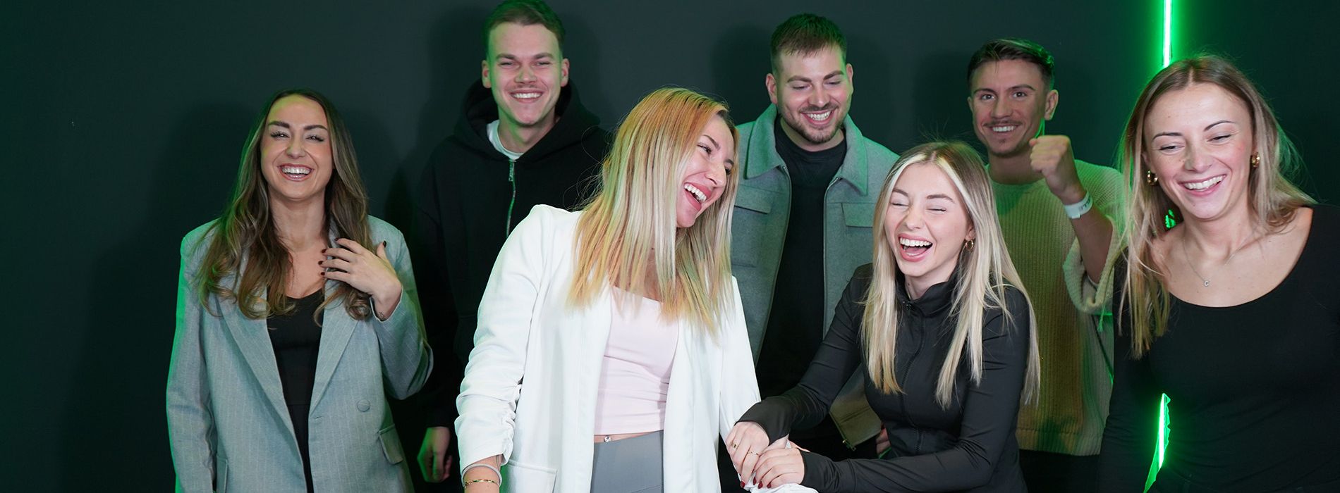 Sieben Personen lachen ausgelassen in Gruppenpose vor schwarzer Wand mit grünem Lichtstreifen.