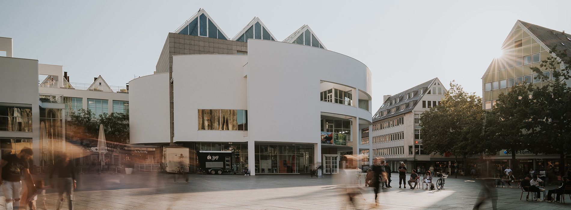 Weitwinkelaufnahme des Stadthauses Ulm mit umliegender urbaner Architektur. Die helle Fassade des Gebäudes hebt sich von der Umgebung ab