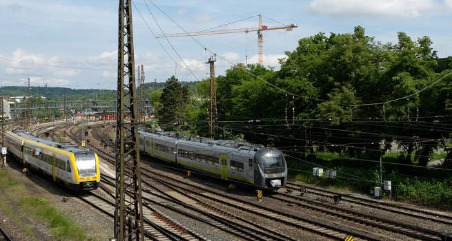 Bahngleise mit mehreren Zügen, darunter ein gelber Nahverkehrszug und ein grauer Zug mit gelben Akzenten. Im Hintergrund sind Baukräne, Gebäude und grüne Bäume zu sehen. Oberleitungen und Strommasten durchziehen die Szene.