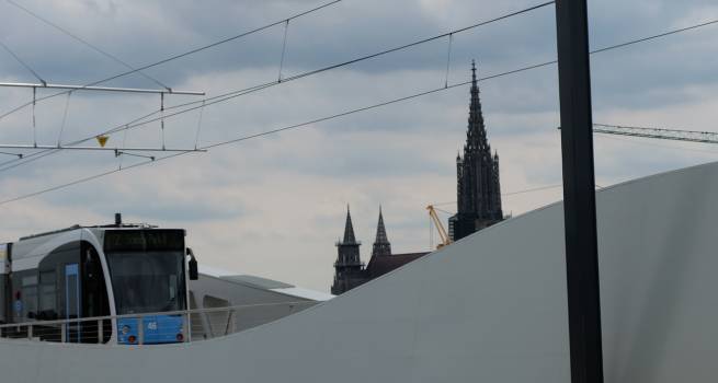 Eine moderne Straßenbahn fährt auf einer erhöhten Trasse mit geschwungenen weißen Wänden. Im Hintergrund ist das Ulmer Münster mit seinem markanten Turm zu sehen, während der Himmel bewölkt ist.