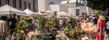Marktbesucher betrachten Pflanzen an einem Verkaufsstand auf belebtem Pflanzenmarkt.