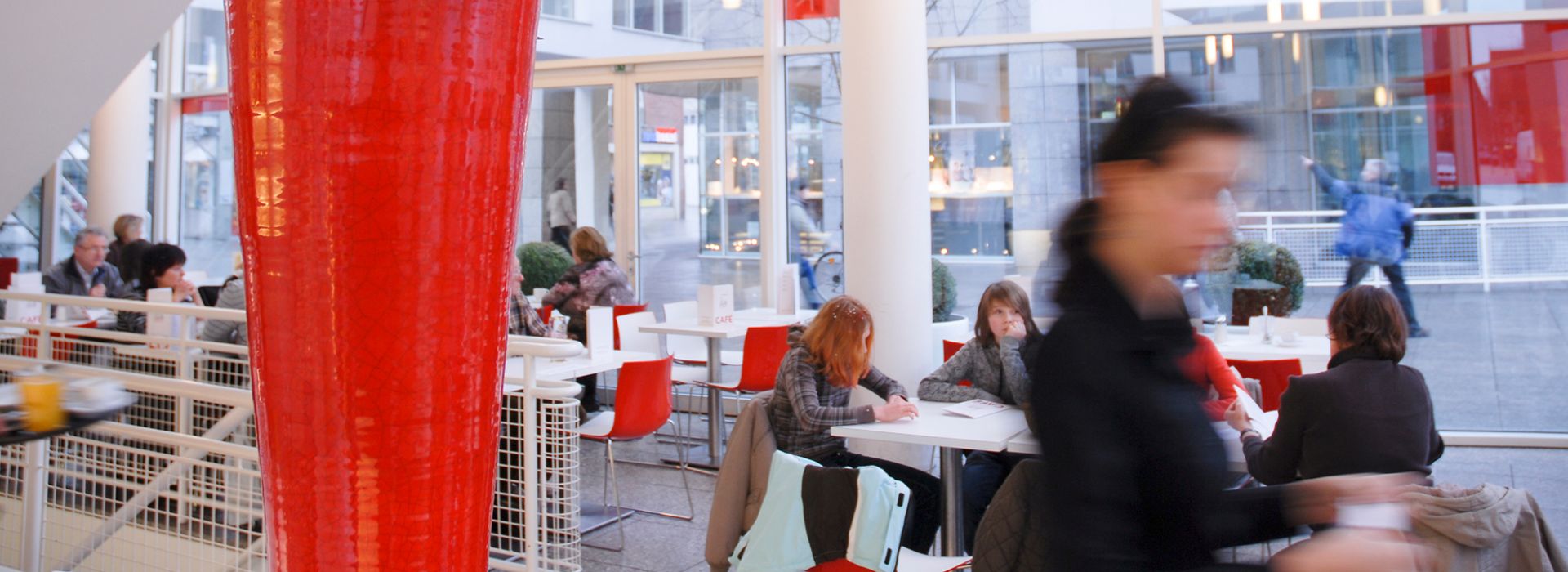 Ein belebtes Café mit großen Fenstern, Menschen sitzen an weißen Tischen mit roten Stühlen, eine Kellnerin serviert.