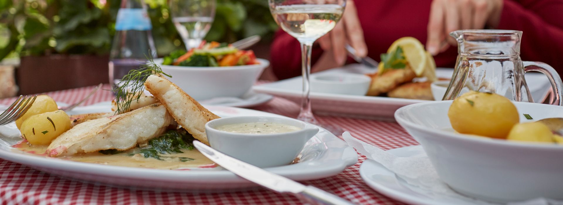 Ein Teller mit frisch zubereitetem Fischfilet auf einer hellen Sauce mit Spinat, serviert mit kleinen Kartoffeln und einer Schale mit Dip. Der Teller steht auf einer rot-weiß karierten Tischdecke, im Hintergrund sind weitere Gerichte, ein Glas Weißwein und eine Person beim Essen zu sehen.