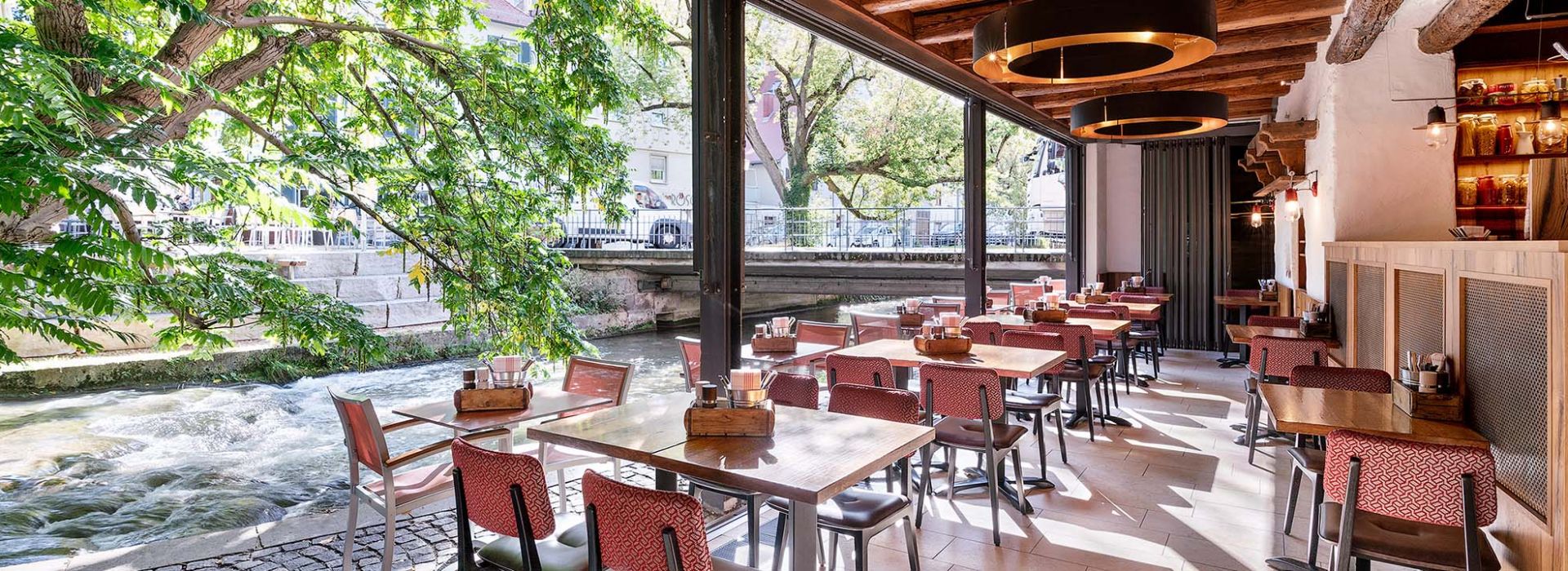 Ein lichtdurchfluteter Innenbereich des Restaurants Tanivera mit großen offenen Fenstern, durch die ein malerischer Bach fließt. Holztische und rot gepolsterte Stühle schaffen eine warme Atmosphäre unter den rustikalen Holzbalken der Decke.