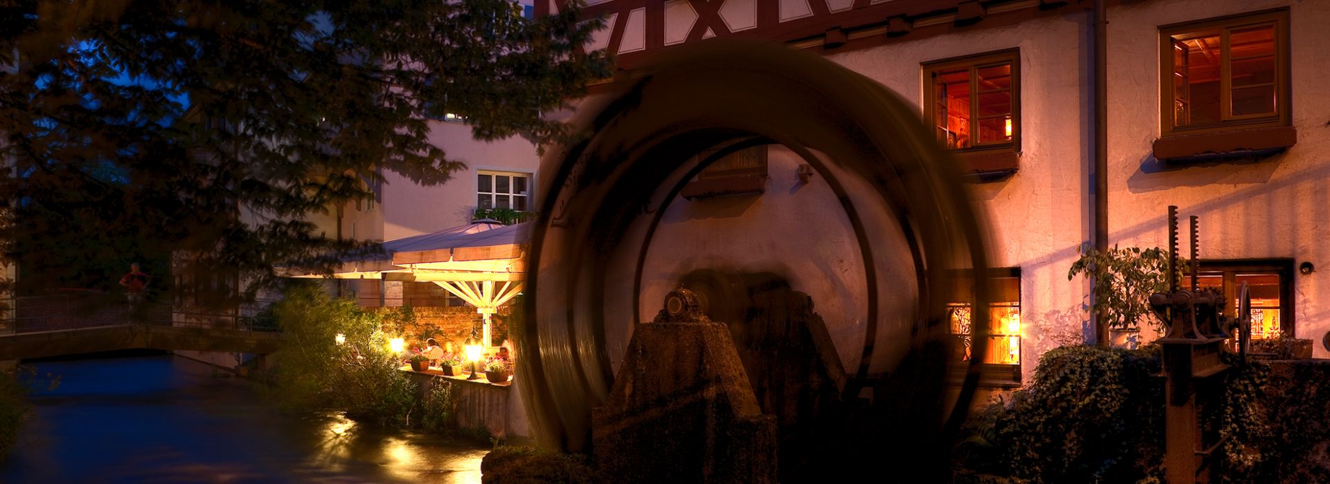 Das große, hölzerne Mühlrad dreht sich in der Dunkelheit vor der beleuchteten Fassade der Lochmühle. Die Fenster des Restaurants strahlen ein warmes Licht aus. Die Außenterrasse ist ebenfalls stimmungsvoll beleuchtet, während sich das Wasser ruhig durch die Szene schlängelt.