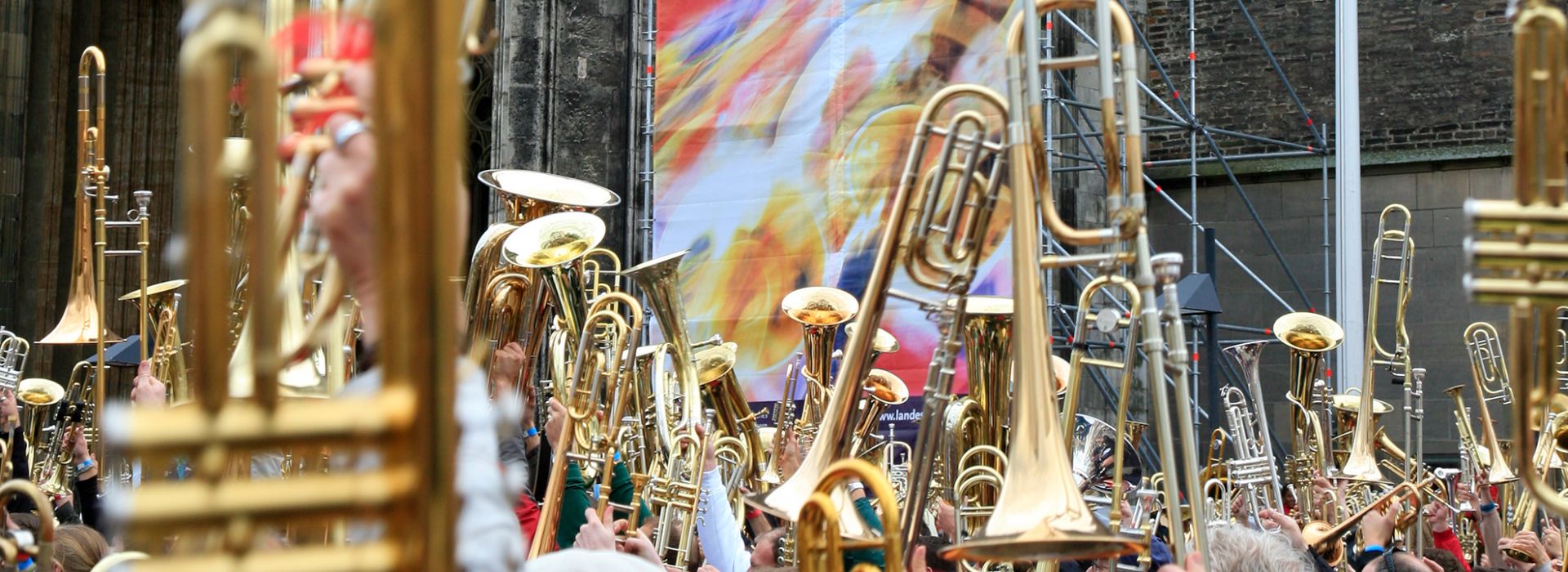 Große Gruppe hebt Blechblasinstrumente in die Luft vor einer Kirchenfassade.