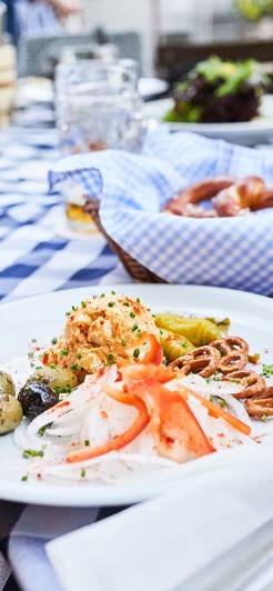 Ein bayerisches Brotzeitbrett mit Obazda, Brezeln, Oliven und Zwiebeln auf einer blau-weißen Tischdecke.