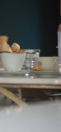 Eine elegante weiße Schale mit goldener Verzierung, gefüllt mit Gebäck, steht auf einem Marmortresen. Dahinter unscharf Gläser, eine Tasse und eine Flasche, die eine gemütliche Café-Atmosphäre schaffen.