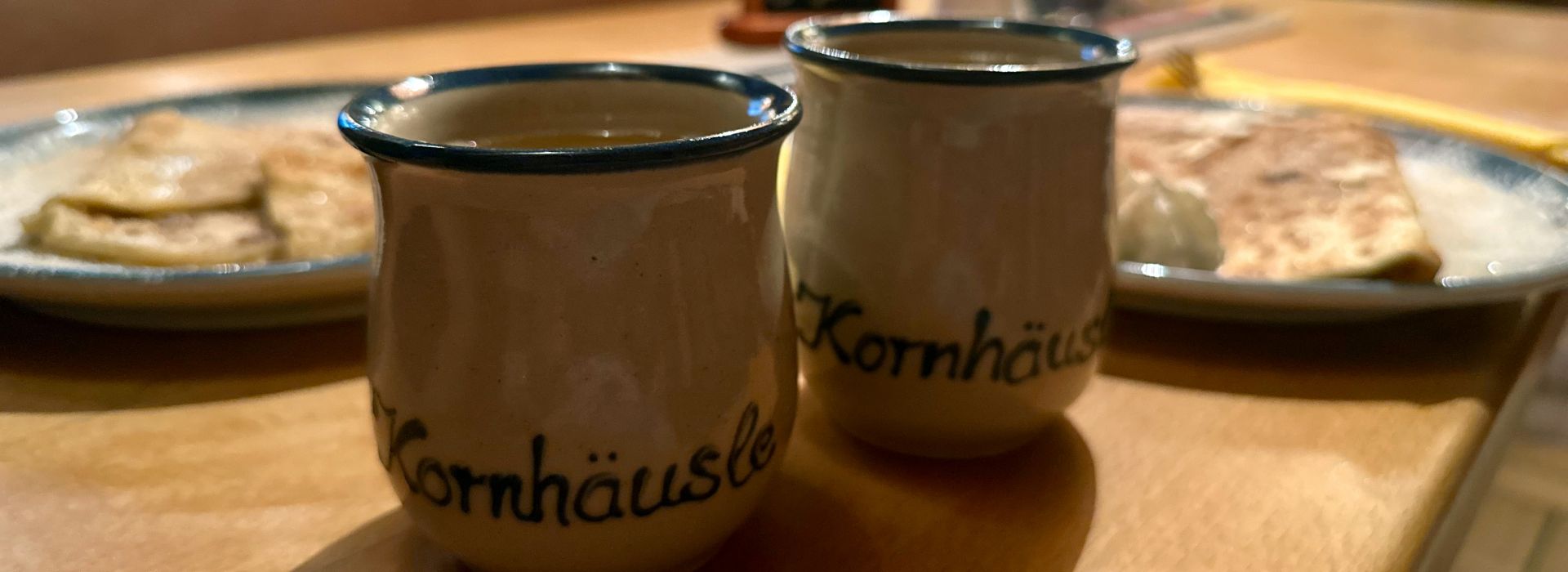  Zwei traditionelle Steinkrüge mit „Kornhäusle“-Schriftzug, gefüllt mit heißem Getränk. Dahinter Teller mit goldbraunen Crêpes, gelbes Serviettenbesteck auf Holztisch.