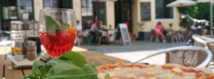 Eine Pizza mit knusprigem Rand und frischem Rucola liegt auf einem Holztisch. Daneben steht ein Glas mit einem roten Spritzgetränk, garniert mit Minzblättern und einer Orangenscheibe. Im Hintergrund ist ein sonniger Außenbereich mit Sitzplätzen und Gästen vor einem gelb gestrichenen Gebäude zu sehen.