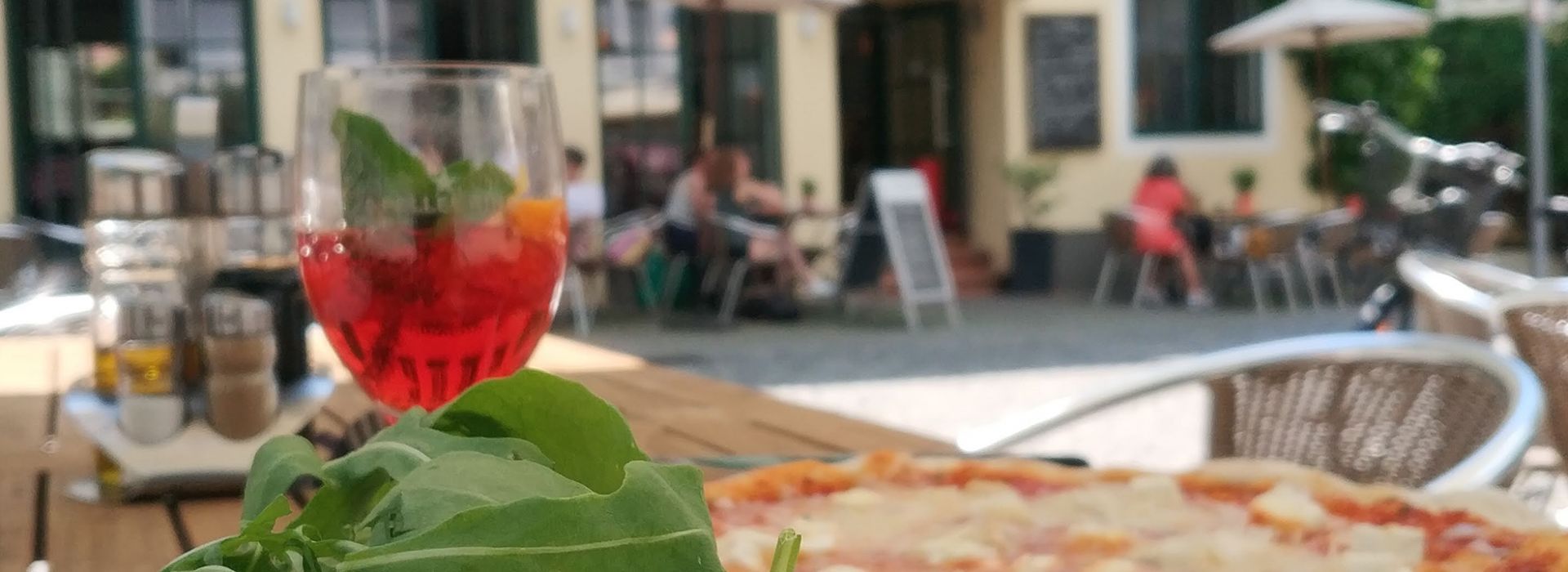 Eine Pizza mit knusprigem Rand und frischem Rucola liegt auf einem Holztisch. Daneben steht ein Glas mit einem roten Spritzgetränk, garniert mit Minzblättern und einer Orangenscheibe. Im Hintergrund ist ein sonniger Außenbereich mit Sitzplätzen und Gästen vor einem gelb gestrichenen Gebäude zu sehen.