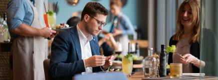 Ein Mann im Anzug und eine Frau im eleganten Outfit genießen ihr Essen im Treibgut. Der Mann konzentriert sich auf sein Gericht, während die Frau lächelnd mit Salat auf der Gabel interagiert. Im Hintergrund serviert das Personal Getränke und Speisen in einer stilvollen Atmosphäre.