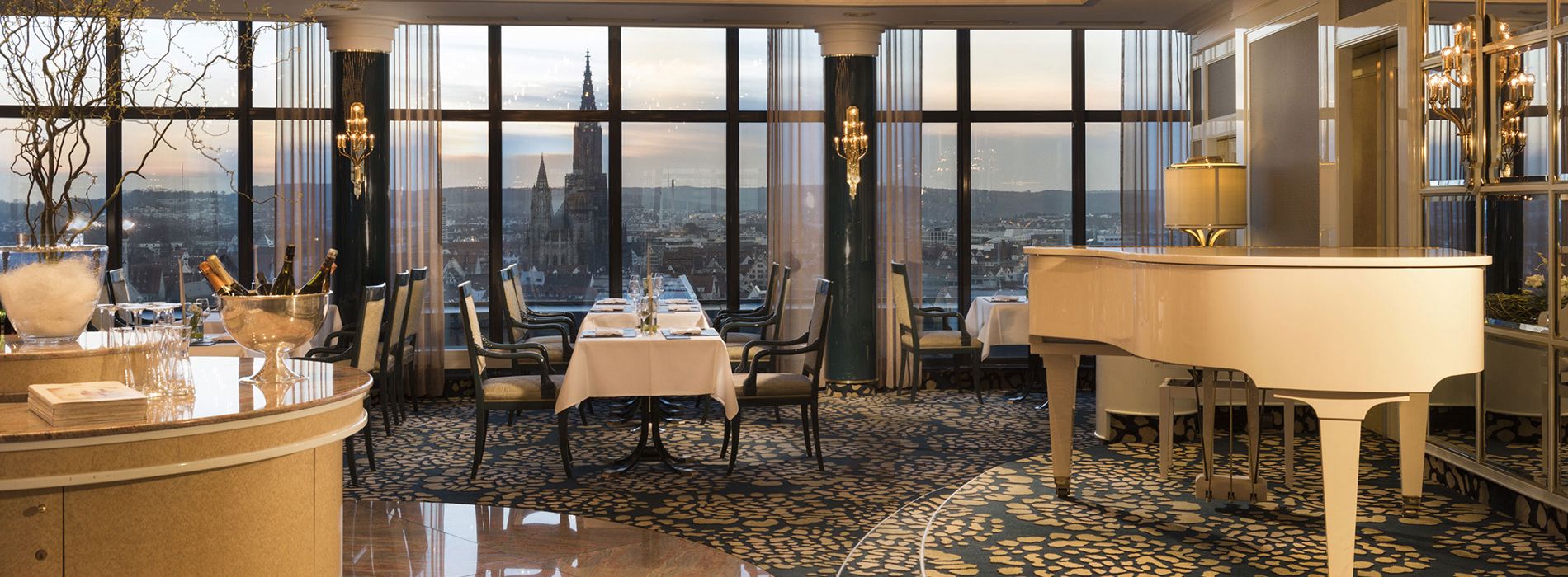 Elegantes Restaurant mit großen Panoramafenstern und Blick auf das Ulmer Münster. Ein weißer Flügel steht im Vordergrund, Tische sind stilvoll eingedeckt.