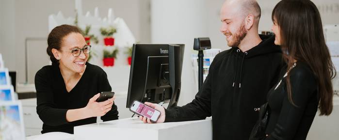 Ein Mann und eine Frau scannen die UlmCard an der Touristeninformation in Ulm. Eine Mitarbeiterin hinter dem Tresen lächelt und hält ebenfalls ein Smartphone.