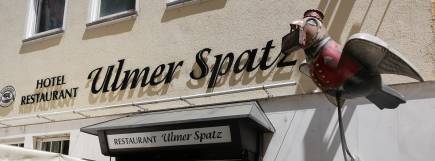 Der Schriftzug „Hotel Restaurant Ulmer Spatz“ an einer beige gestrichenen Hauswand, darüber eine dekorative Metallfigur eines Spatzen.