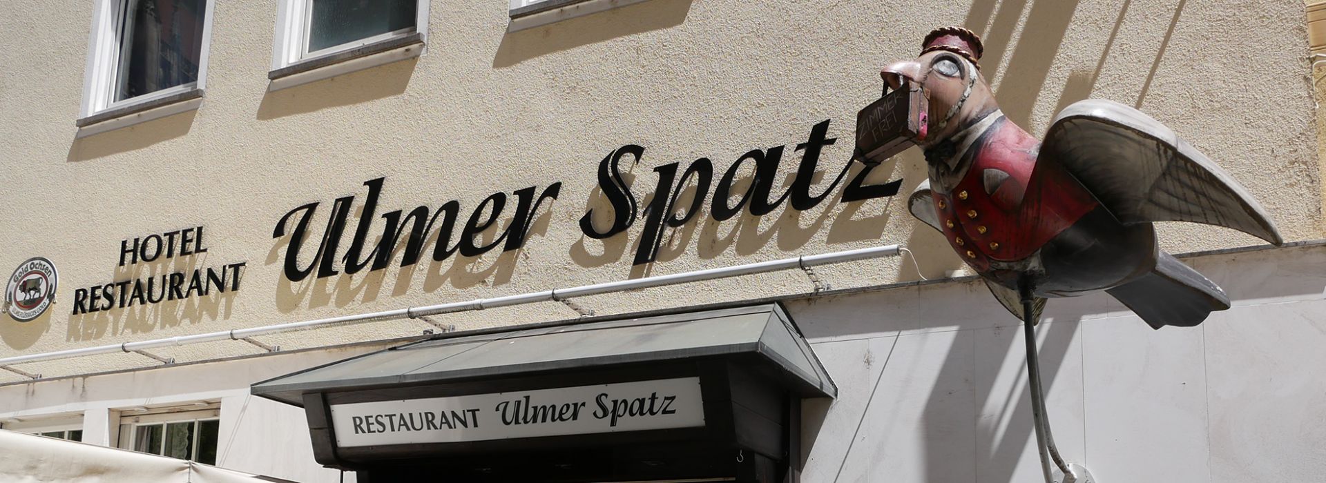 Der Schriftzug „Hotel Restaurant Ulmer Spatz“ an einer beige gestrichenen Hauswand, darüber eine dekorative Metallfigur eines Spatzen.