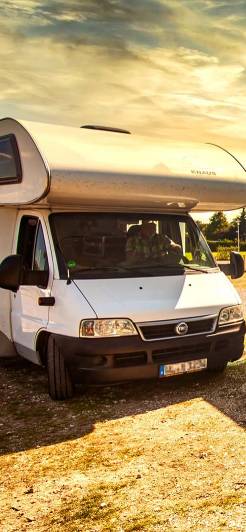 Ein weißes KNAUS-Wohnmobil steht auf einem Stellplatz in der Abendsonne. Der Fahrer sitzt im Fahrzeug und schaut hinaus. Im Hintergrund sind Bäume und Lampen zu sehen, die den Stellplatz begrenzen.