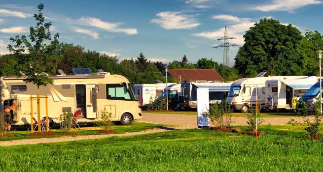 Wohnmobilstellplatz mit mehreren Fahrzeugen, die auf gepflegten Parzellen stehen. Im Vordergrund ein beige Wohnmobil mit Markise und Campingstühlen. Die Umgebung ist grün und gepflegt, mit Bäumen und Blumenbeeten. Im Hintergrund sind weitere Wohnmobile sowie ein Strommast zu sehen.