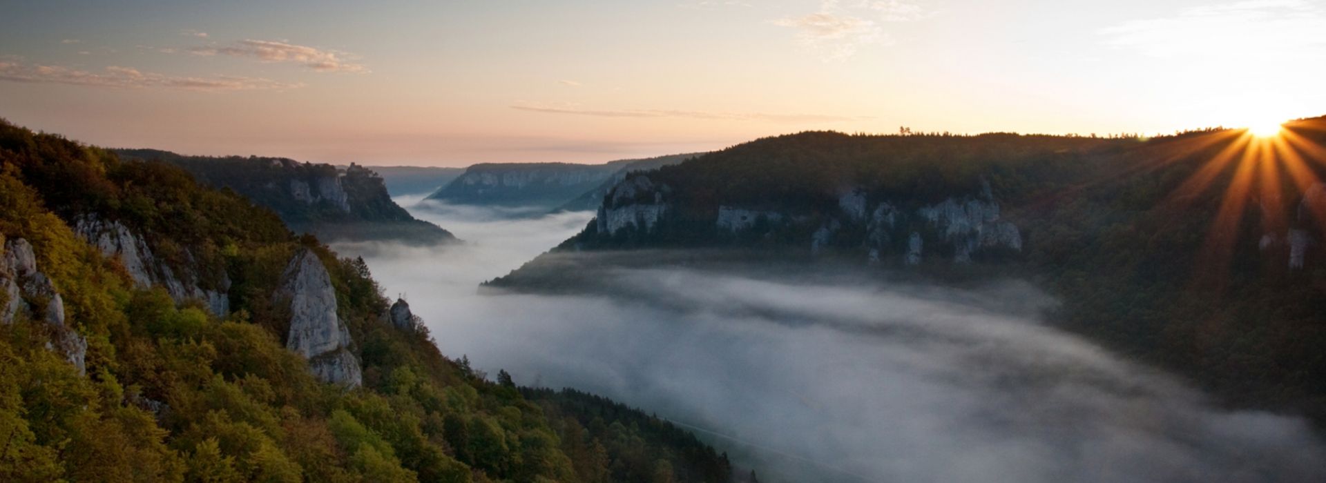 Panoramablick auf nebelverhangenes Flusstal im Morgenlicht mit bewaldeten Höhenzügen.