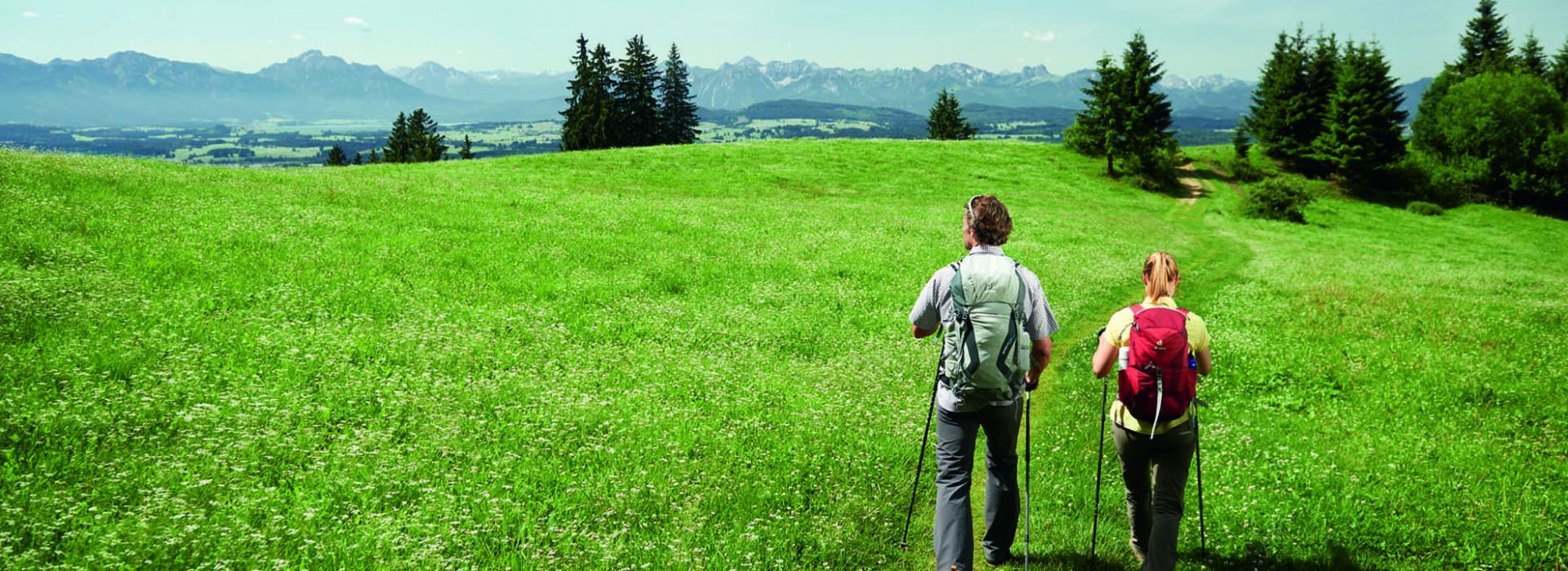 Zwei Personen wandern durch offene grüne Landschaft mit Blick auf entfernte Berge.
