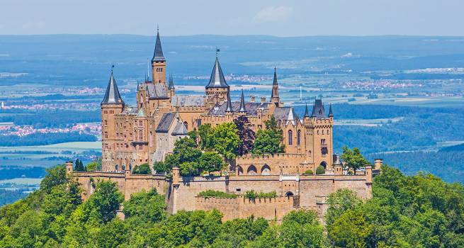 Die beeindruckende Burg Hohenzollern thront majestätisch auf einem bewaldeten Hügel, mit Blick auf eine weitläufige Landschaft und entfernte Dörfer