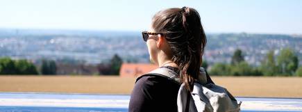 Eine Frau mit Rucksack und Sonnenbrille blickt von einer Aussichtstafel aus auf die Stadt Ulm und die umliegende Landschaft