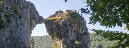 Felsformation mit natürlicher Steinbrücke über Tal, umgeben von bewaldeter Landschaft.
