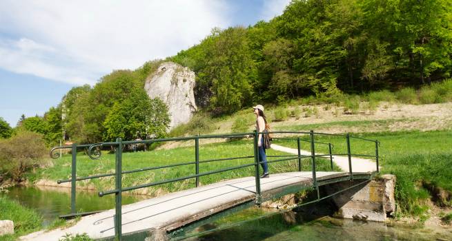 Eine Frau mit Kappe und Rucksack steht auf einer kleinen Brücke über einen Bach, umgeben von grüner Natur und Felsen