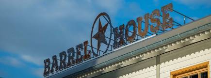 Ein rostiges Metallschild mit der Aufschrift „Barrel House“ auf dem Dach des Gebäudes, mit einem großen Sternsymbol daneben. Der Himmel im Hintergrund ist blau mit vereinzelten Wolken.