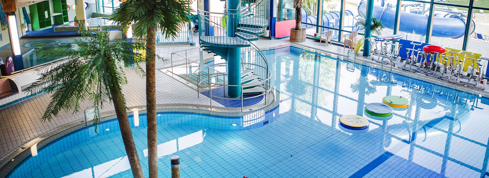 Modernes Hallenbad mit Rutsche, Wasserbecken und großen Fensterflächen.