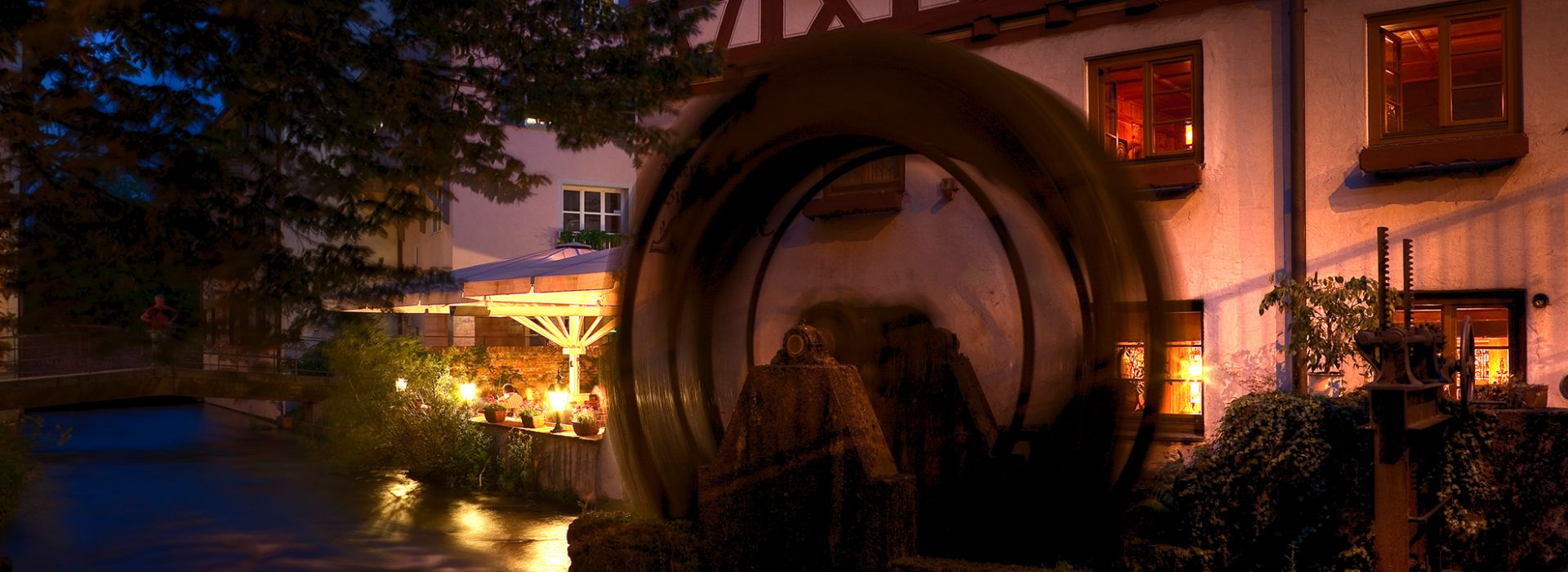 Beleuchtetes Wasserrad an historischem Gebäude bei Nacht mit Spiegelung im Wasser.