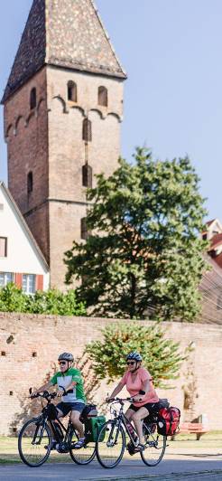 Mehrere Radfahrer mit Helmen fahren entlang der Stadtmauer in Ulm. Im Hintergrund ist ein historischer Turm mit farbigem Dach sowie Fachwerkhäuser zu sehen.