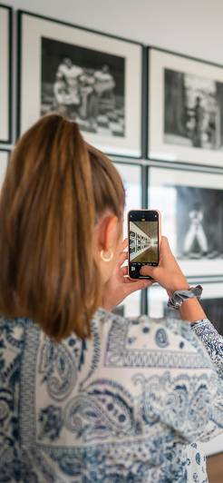 Eine junge Frau mit Pferdeschwanz und einem blauen Paisley-Hemd fotografiert mit ihrem Smartphone eine Wand mit Schwarz-Weiß-Fotografien in einer Kunstausstellung