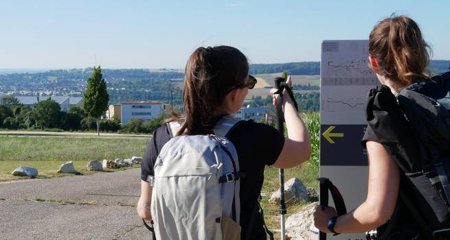 Zwei Frauen mit Rucksäcken und Wanderstöcken stehen an einem Wegweiser mit Höhenprofil und Richtungspfeil auf einem sonnigen Wanderweg