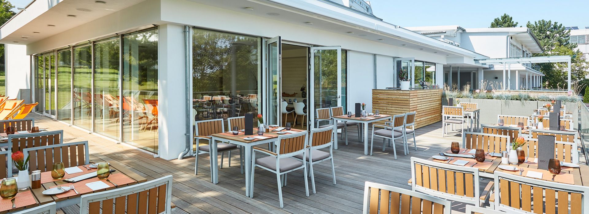 Eine großzügige Außenterrasse mit modernen Holztischen und Stühlen, die stilvoll eingedeckt sind. Große Glasfronten verbinden das helle Innere mit der Natur. Im Hintergrund sind Liegestühle und eine grüne Parklandschaft zu sehen