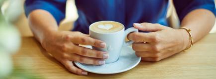 Kaffeegenuss: Frau mit dunkelblauer Bluse hält eine weiße Tasse Cappuccino mit Herz-Latte-Art in den Händen, entspannte Atmosphäre im Café.