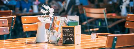 Ein einladend gedeckter Holztisch im Biergarten mit Blume in einer weißen Vase, Speisekarten und einer Kreidetafel mit der Aufschrift „Herzlich willkommen“.