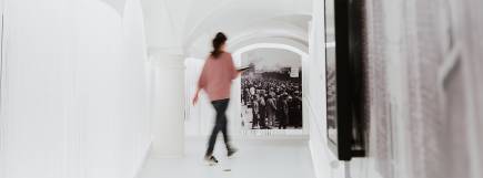 Ein heller Gang mit weißen Wänden und einer gewölbten Decke. Eine Frau in einem rosa Pullover und Jeans geht mit einem Tablet in der Hand durch den Flur. Im Hintergrund ist ein großes historisches Foto von Menschen in einem Konzentrationslager zu sehen.