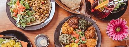 Eine bunte Auswahl an äthiopischen Gerichten auf schwarzen Tellern, darunter injera (traditionelles Fladenbrot), Gemüse, Linsen, Salate und verschiedene würzige Beilagen. Daneben ein Glas mit rotem Getränk, dekoriert mit Orangen- und Grapefruit-Scheiben und schwarz-weißen Strohhalmen. Eine brennende Kerze sorgt für eine gemütliche Atmosphäre.