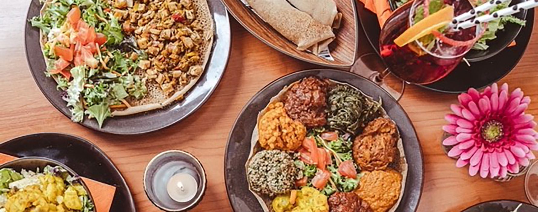 Eine bunte Auswahl an äthiopischen Gerichten auf schwarzen Tellern, darunter injera (traditionelles Fladenbrot), Gemüse, Linsen, Salate und verschiedene würzige Beilagen. Daneben ein Glas mit rotem Getränk, dekoriert mit Orangen- und Grapefruit-Scheiben und schwarz-weißen Strohhalmen. Eine brennende Kerze sorgt für eine gemütliche Atmosphäre.